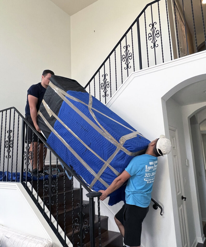Two Orlando Express Movers carrying a wrapped piece of furniture down a staircase